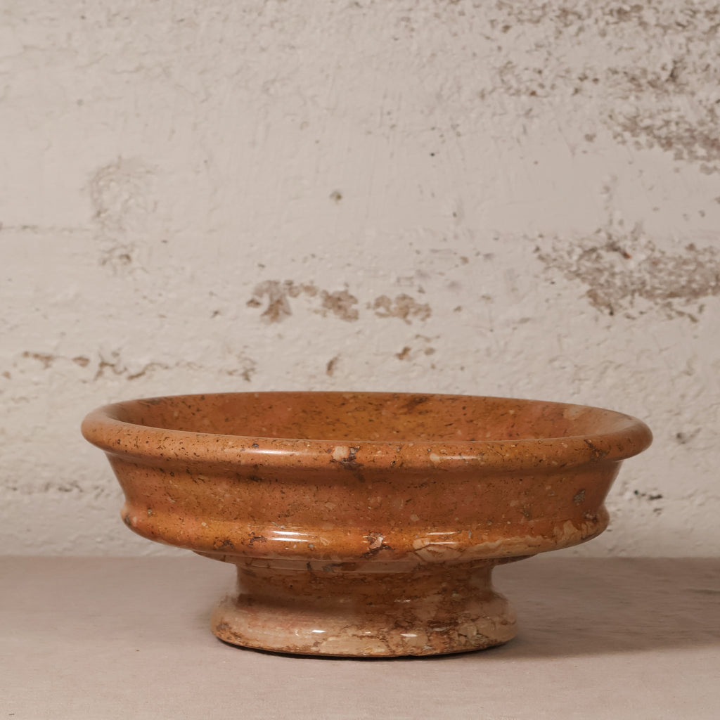 A rustic terracotta clay pedestal bowl with a textured, earthy orange-brown glaze, set against a distressed off-white plaster wall.