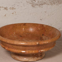 A rustic terracotta-colored ceramic bowl with a raised pedestal base, featuring a speckled glaze and a weathered finish, set against a textured off-white wall.