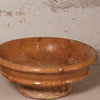 A rustic terracotta-colored ceramic bowl with a raised pedestal base, featuring a speckled glaze and a weathered finish, set against a textured off-white wall.
