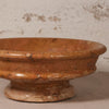 A terracotta-colored stone pedestal bowl with a textured, speckled finish, shown close-up against a rustic, light-colored plaster wall.