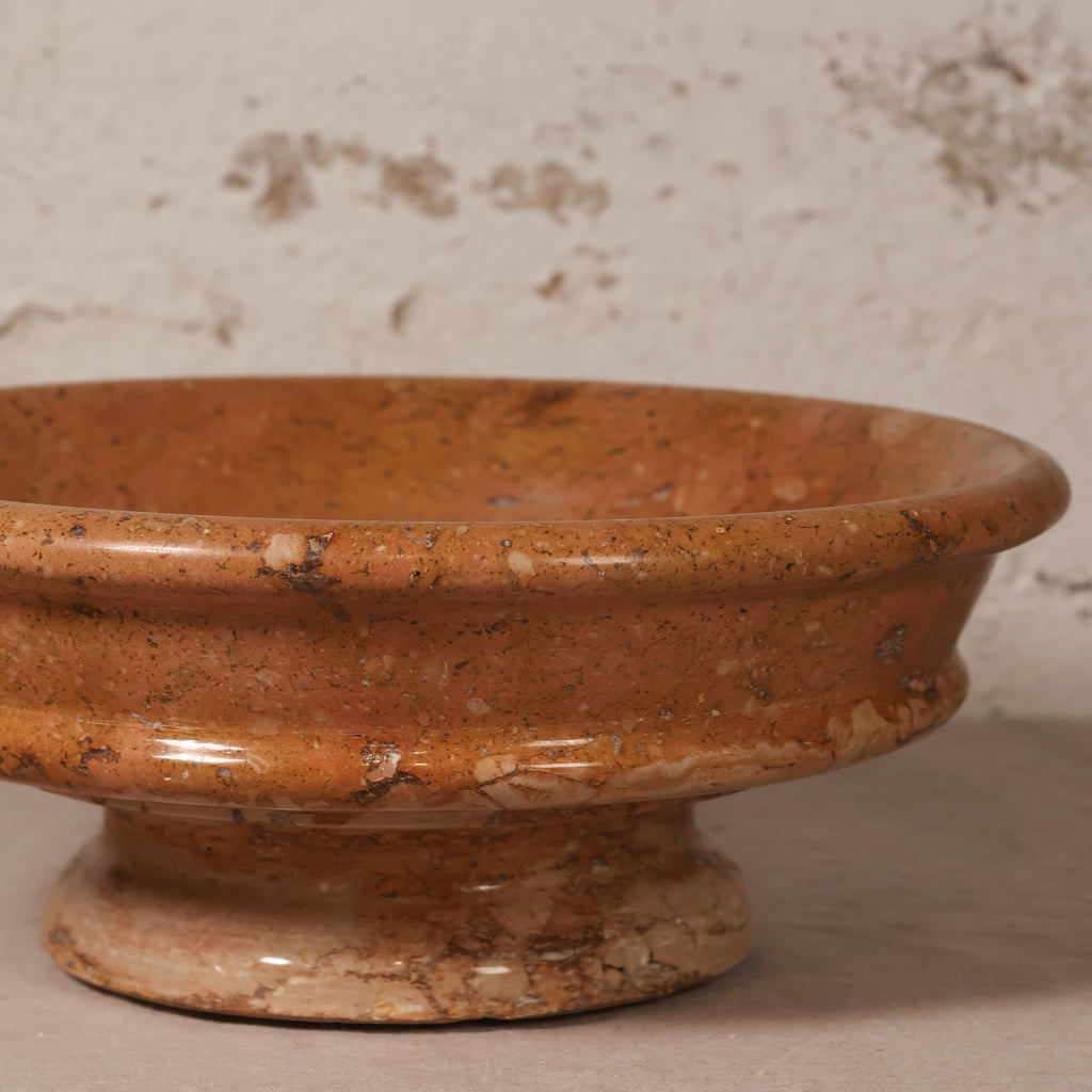 A terracotta-colored stone pedestal bowl with a textured, speckled finish, shown close-up against a rustic, light-colored plaster wall.