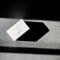 A rectangular white bar of soap with embossed text and a small row of dots, photographed on a textured gray surface with dramatic diagonal shadows against a black background.