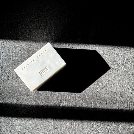 A rectangular white bar of soap with embossed text and a small row of dots, photographed on a textured gray surface with dramatic diagonal shadows against a black background.