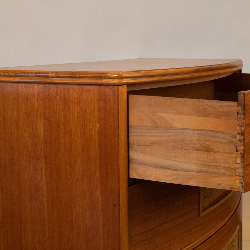 Close-up of a warm medium-brown wooden cabinet with a curved top and an open drawer showing dovetail joint construction.
