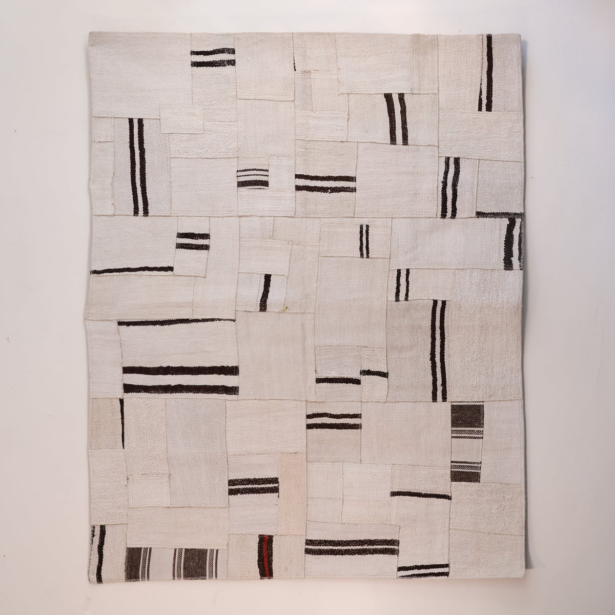 A square textile wall hanging or rug made from stitched off-white fabric panels with irregular black and dark brown stripes, shown against a plain light wall.