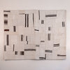 A square textile wall hanging or rug made from stitched off-white fabric panels with irregular black and dark brown stripes, shown against a plain light wall.