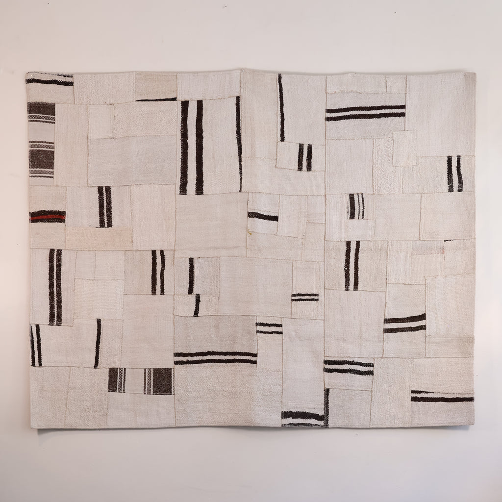 A square textile wall hanging or rug made from stitched off-white fabric panels with irregular black and dark brown stripes, shown against a plain light wall.