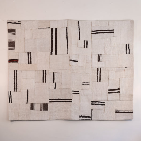 A square textile wall hanging or rug made from stitched off-white fabric panels with irregular black and dark brown stripes, shown against a plain light wall.