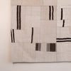 A close-up of a woven textile or rug with a patchwork design in warm off-white and beige, accented by dark brown and black vertical stripe patterns.