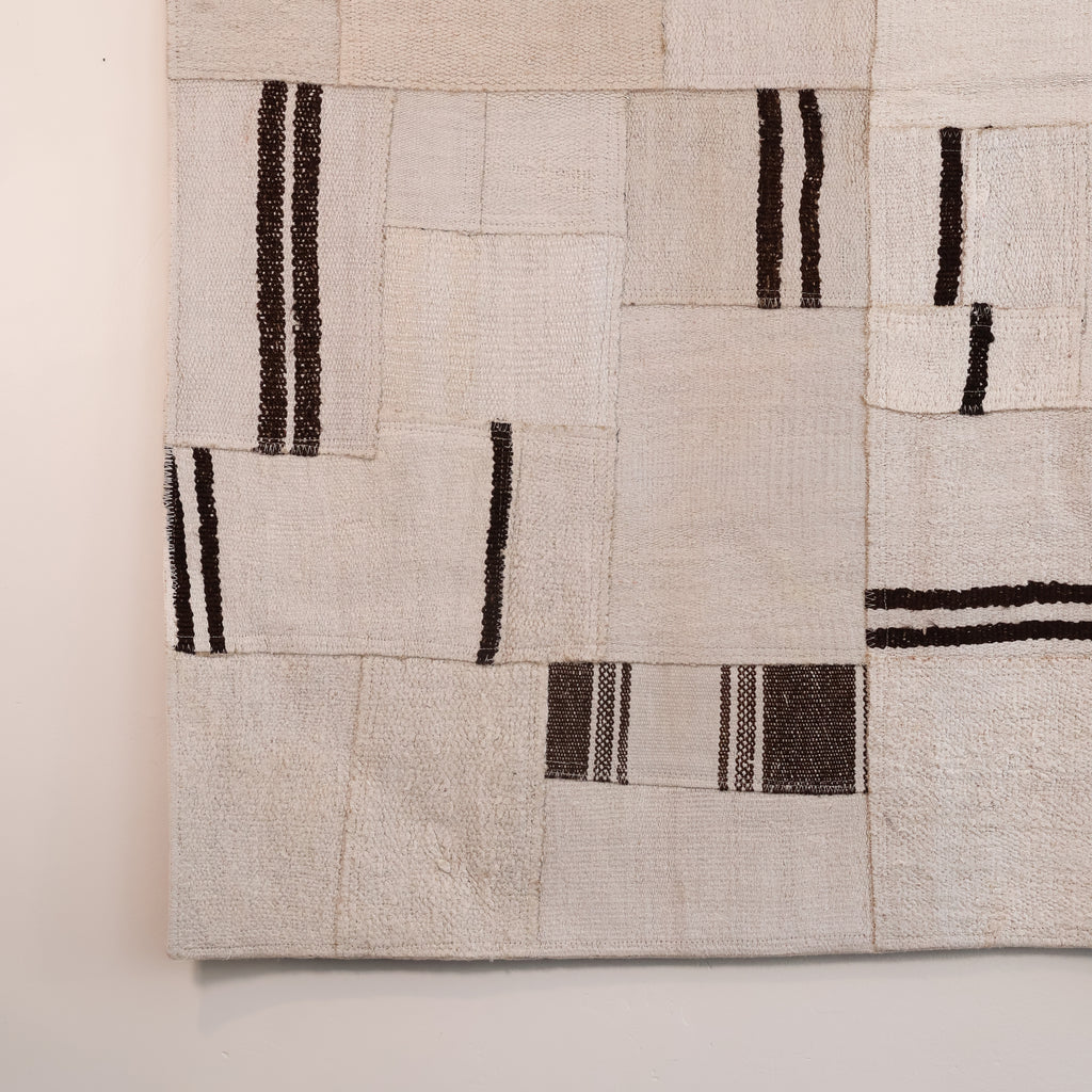A close-up of a woven textile or rug with a patchwork design in warm off-white and beige, accented by dark brown and black vertical stripe patterns.