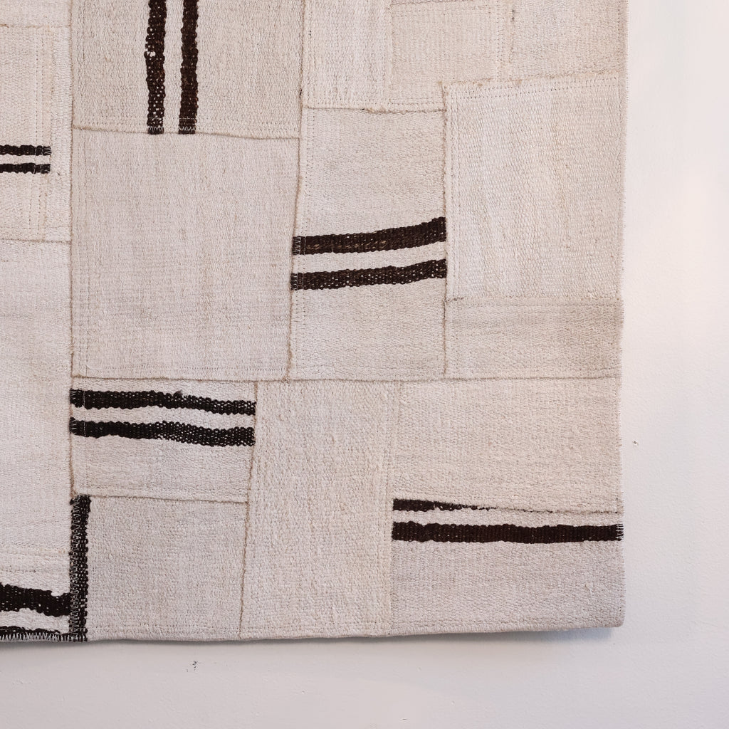 A close-up of a woven textile or rug with a patchwork-style geometric pattern in off-white and beige with dark brown stripes, shown against a plain white background.
