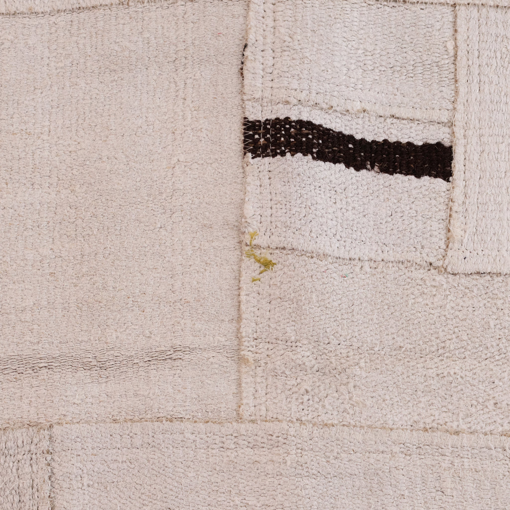 A close-up of a textured woven rug or textile in warm off-white/cream with a dark brown horizontal stripe and a small yellow-green accent near the center.