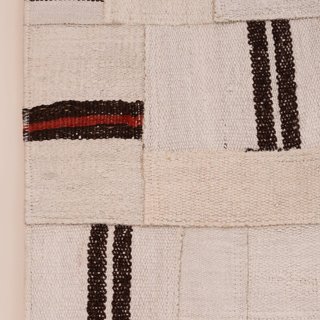 A close-up of a woven textile or rug showing a patchwork of cream and off-white woven squares with dark brown stripes and one horizontal stripe in red.