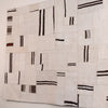 A square textile wall hanging or rug with an abstract patchwork design of cream and off-white rectangular sections, accented by vertical and horizontal black stripes.
