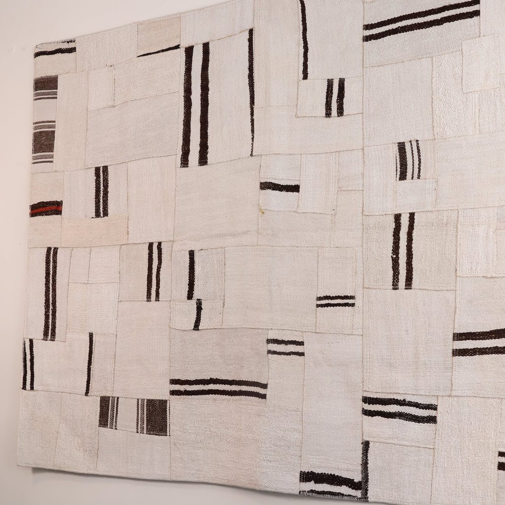 A square textile wall hanging or rug with an abstract patchwork design of cream and off-white rectangular sections, accented by vertical and horizontal black stripes.
