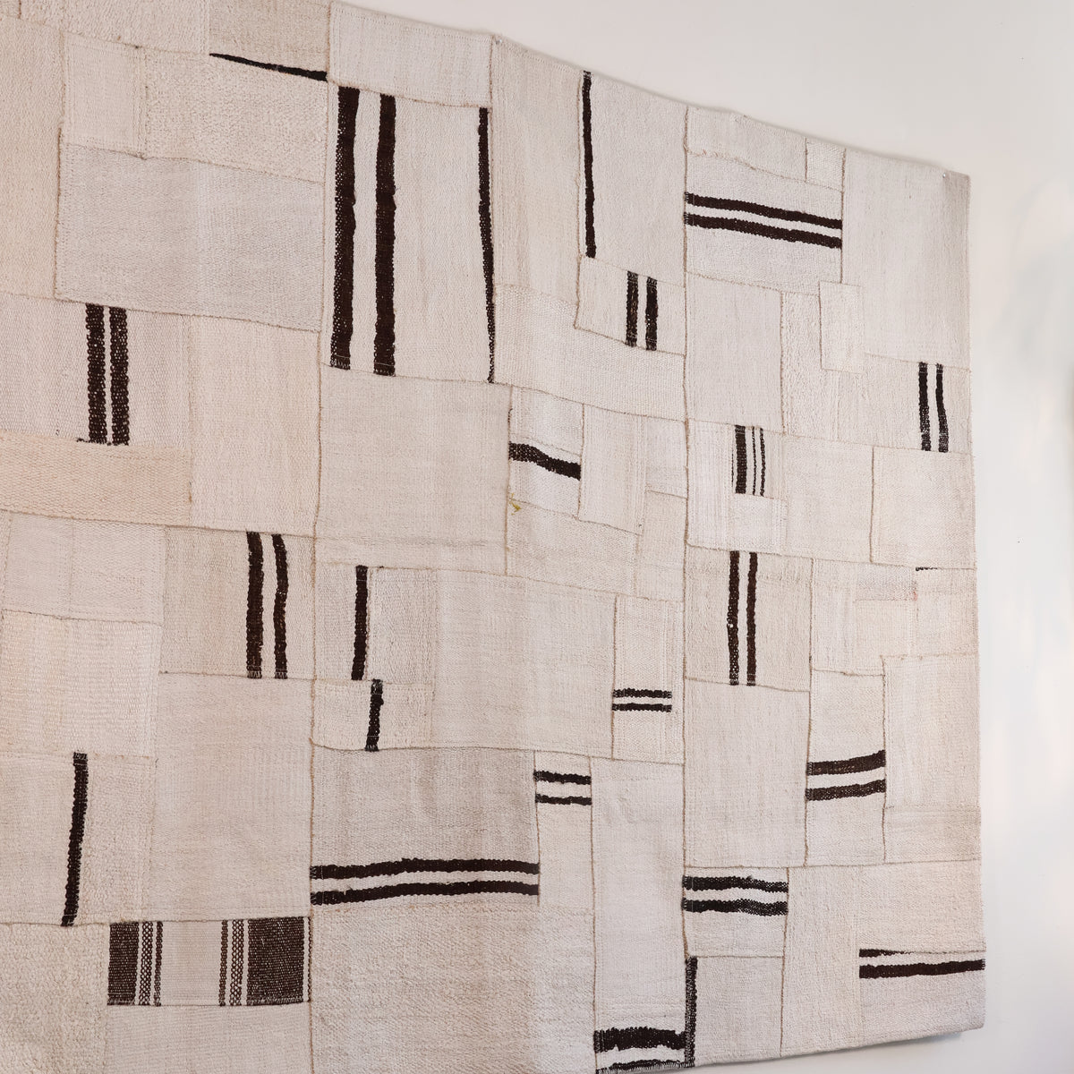 A large rectangular textile wall hanging or rug made from overlapping off-white woven fabric panels with irregular black stripe patterns, shown against a plain white wall.
