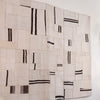 A large rectangular textile wall hanging or rug made from overlapping off-white woven fabric panels with irregular black stripe patterns, shown against a plain white wall.