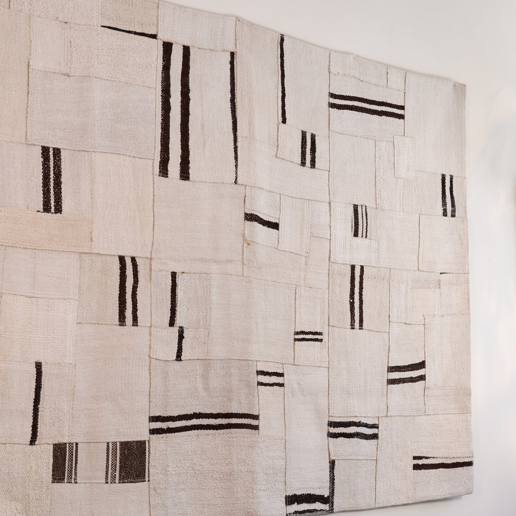 A large rectangular textile wall hanging or rug made from overlapping off-white woven fabric panels with irregular black stripe patterns, shown against a plain white wall.