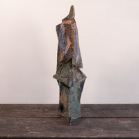 A rustic, abstract hand-carved wooden sculpture with a weathered, distressed finish in shades of brown, green, and blue, standing on a dark wooden surface against a plain off-white wall.