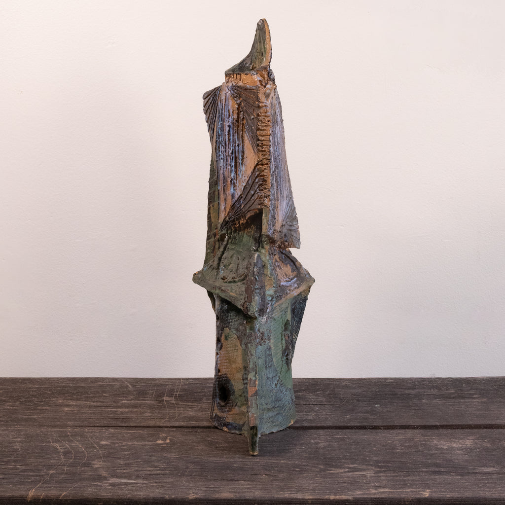 A rustic, abstract hand-carved wooden sculpture with a weathered, distressed finish in shades of brown, green, and blue, standing on a dark wooden surface against a plain off-white wall.