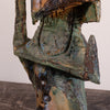 Close-up of a textured abstract sculpture with layered metallic and earthy tones, including green, bronze, brown, and black, set against a plain light background.