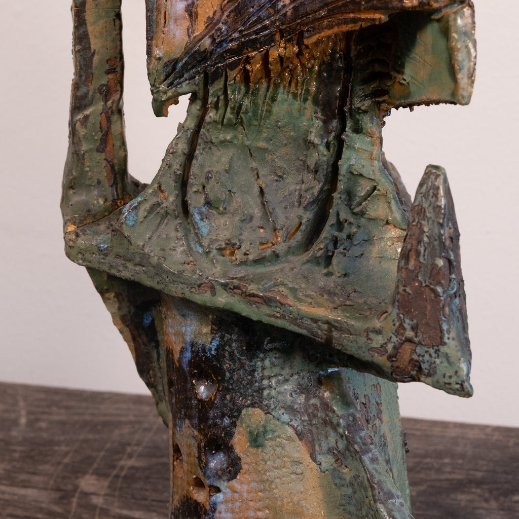 Close-up of a textured abstract sculpture with layered metallic and earthy tones, including green, bronze, brown, and black, set against a plain light background.