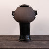 A dark brown and black glazed ceramic lidded urn with side handles, displayed on a rustic dark wooden surface against a plain off-white wall.