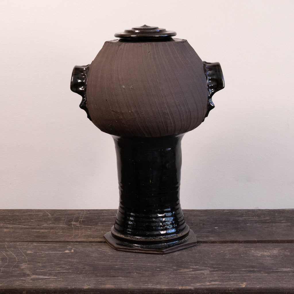 A dark brown and black glazed ceramic lidded urn with side handles, displayed on a rustic dark wooden surface against a plain off-white wall.