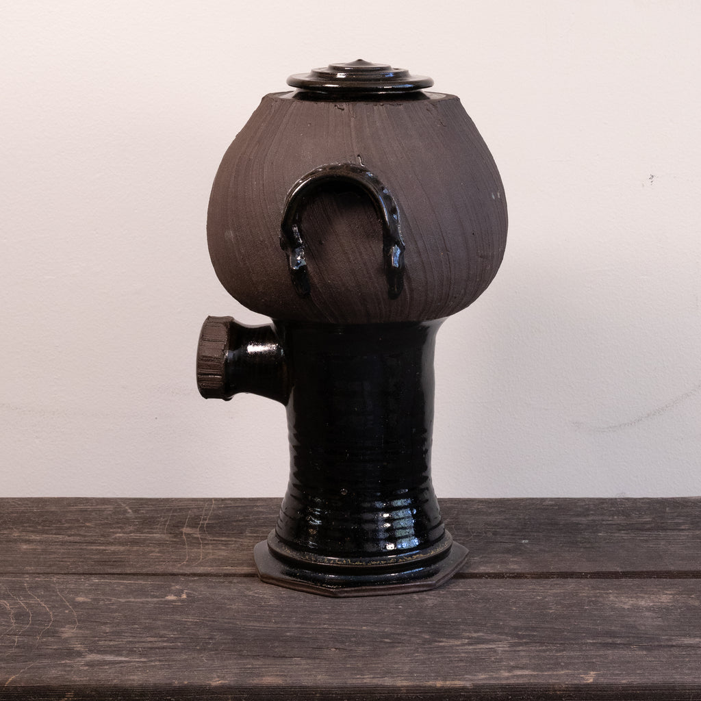 A handmade dark brown and black glazed ceramic vessel with a rounded bulbous top, a side protruding spout, and a small lid on top, displayed on a rustic wooden surface against a plain off-white wall.