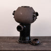 A dark glazed ceramic vessel with a rounded bulbous top, side protruding openings, and a separate matching lid placed beside it on a wooden surface against a plain light wall.