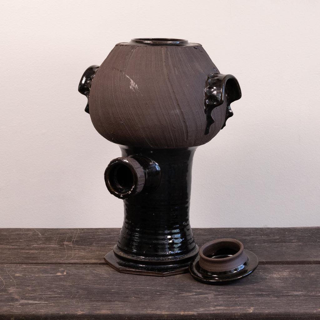 A dark glazed ceramic vessel with a rounded bulbous top, side protruding openings, and a separate matching lid placed beside it on a wooden surface against a plain light wall.