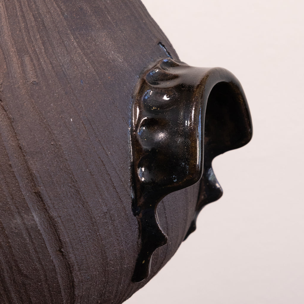 Close-up of a dark brown textured ceramic vessel with a glossy black dripping glaze detail on a light off-white background.