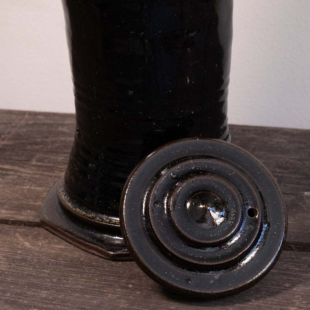 A close-up of a glossy black ceramic vessel or container with a matching round lid placed in front, resting on a dark wooden surface against a light neutral background.