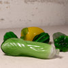 A set of hand-blown glass vegetables, including a prominent green cabbage in the foreground, a yellow pepper in the center, and other green vegetables in the background, arranged on a textured light beige surface against a rustic white wall.