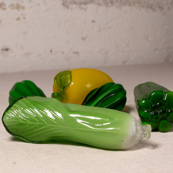 A set of hand-blown glass vegetables, including a prominent green cabbage in the foreground, a yellow pepper in the center, and other green vegetables in the background, arranged on a textured light beige surface against a rustic white wall.