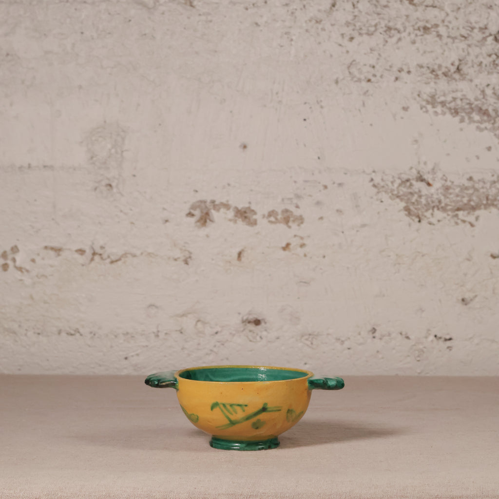 Yellow ceramic bowl with green interior and handles on a textured surface with a beige background