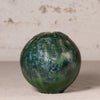 A round ceramic vase with a glossy, marbled green and blue pattern, set against a neutral textured beige background.