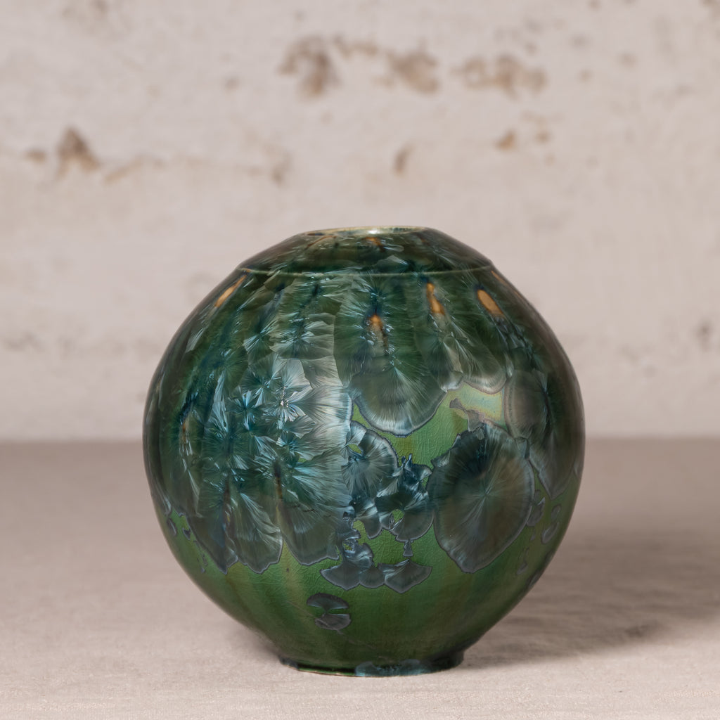 A round ceramic vase with a glossy, marbled green and blue pattern, set against a neutral textured beige background.