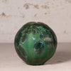 A round green glass vase with a textured, organic leaf-like pattern, set against a neutral beige background.