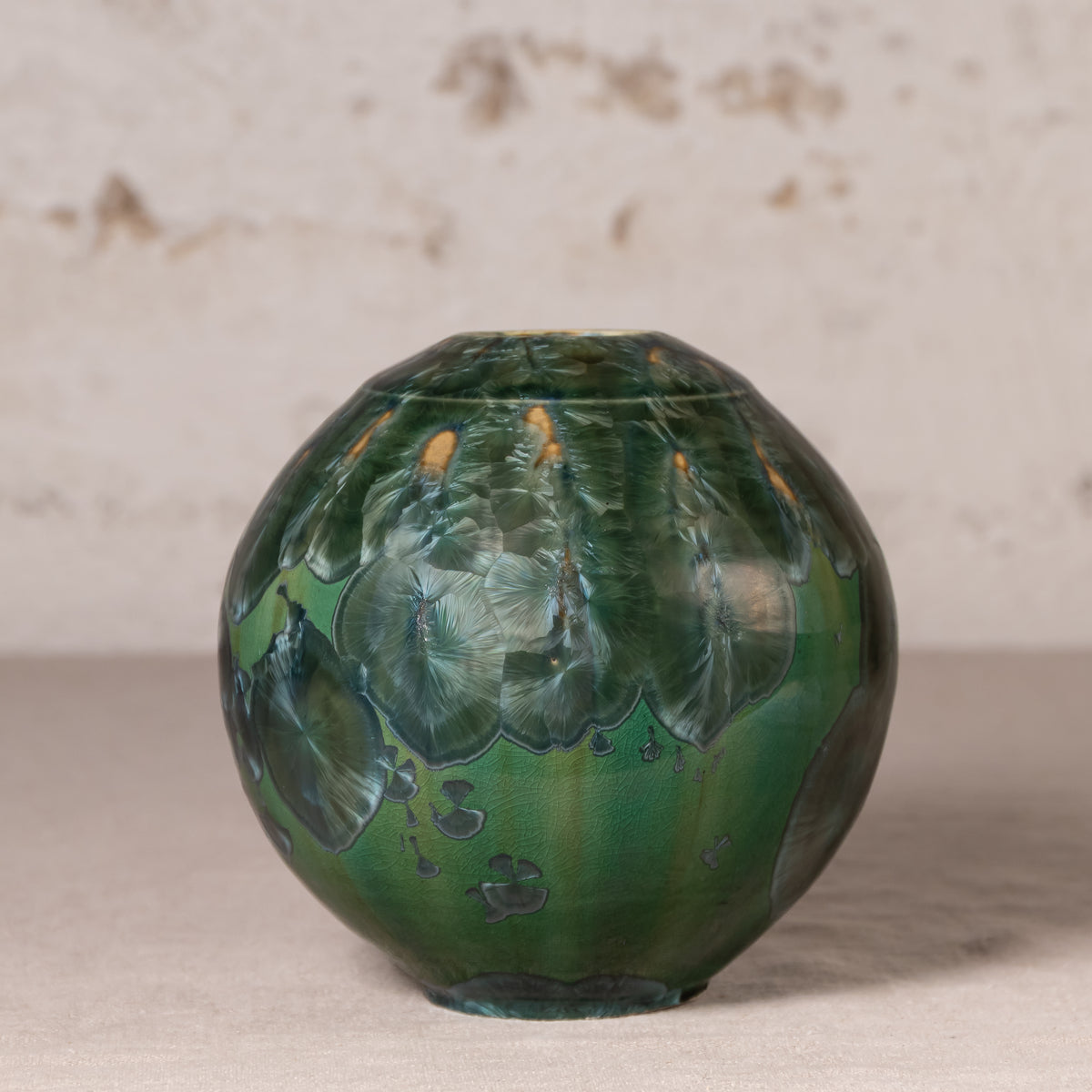 A round ceramic vase with a glossy green and blue marbled pattern, featuring small orange accents near the top, set against a neutral textured background.