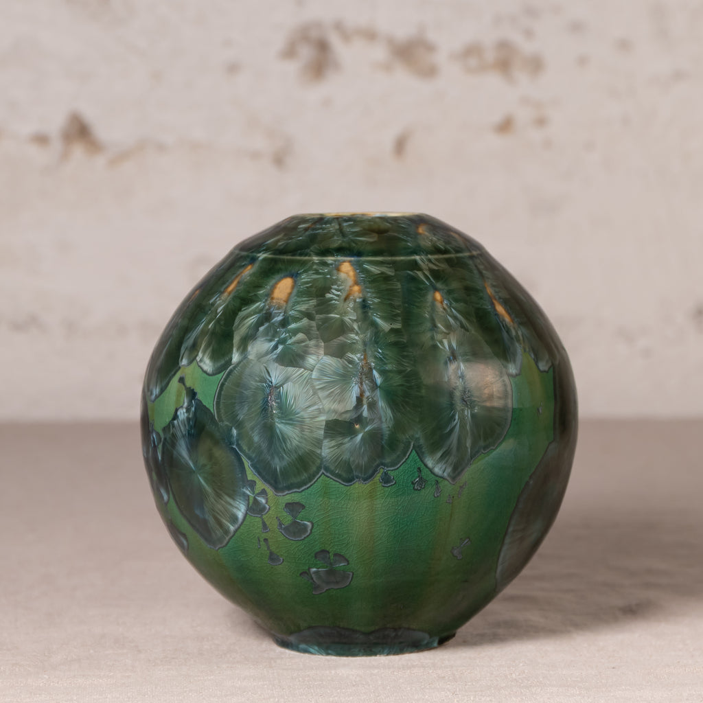 A round ceramic vase with a glossy green and blue marbled pattern, featuring small orange accents near the top, set against a neutral textured background.