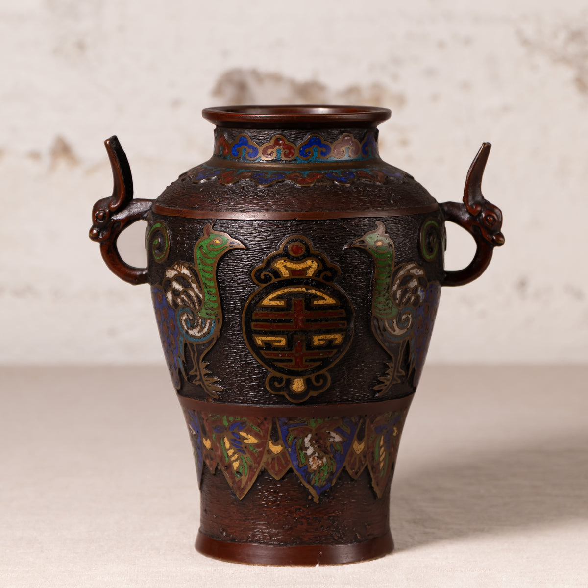 An antique-style Japanese cloisonné bronze vase with dragon handles and phoenix motifs, displayed on a light beige surface against a textured off-white background.