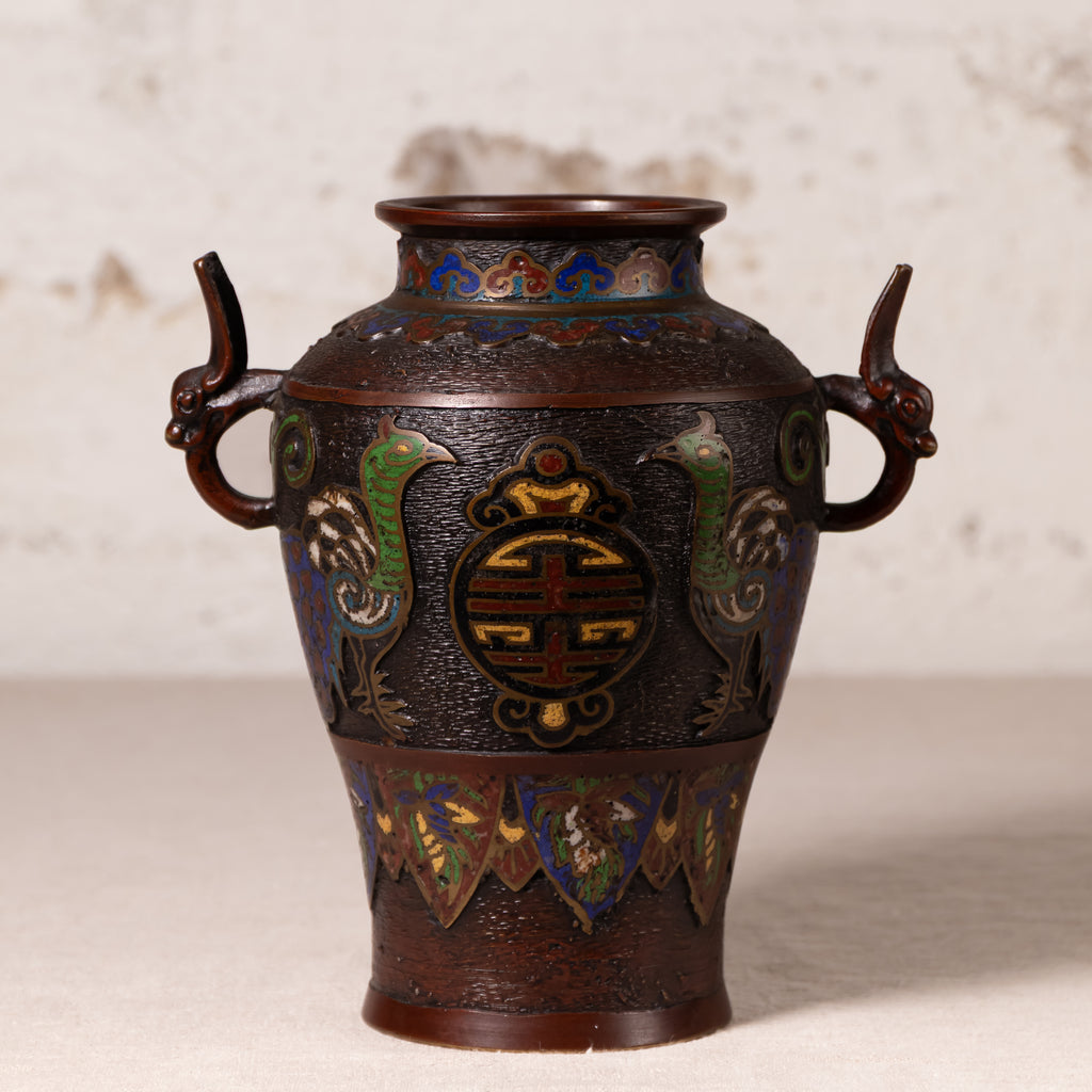 An antique-style Japanese cloisonné bronze vase with dragon handles and phoenix motifs, displayed on a light beige surface against a textured off-white background.