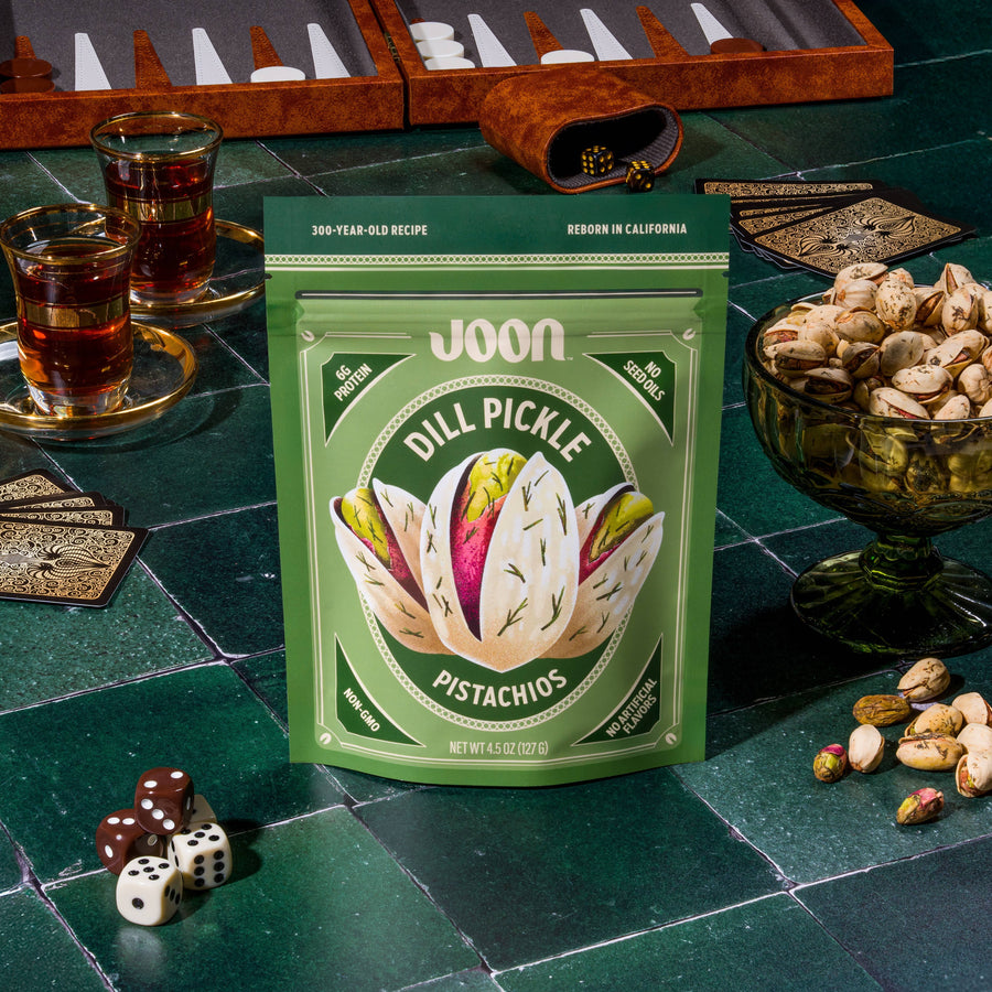 Packaged joon dill pickle pistachios with a green label on a table with dice and tea glasses.