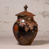 A handmade ceramic lidded jar with a glossy finish, featuring a dark brown and black base with coppery orange and reddish-brown mottled patterns, set against a textured light gray wall.
