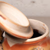 Close-up of a ceramic lidded container with the lid propped open, showing a warm orange-brown glazed exterior and a cream interior.