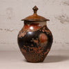 A handmade ceramic lidded jar with a glossy, mottled finish in black, brown, and terracotta orange, set against a textured light gray background.