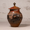 A handmade ceramic lidded jar with a rounded body, featuring a glossy brown and black mottled glaze with terracotta orange patches, set against a textured light gray wall.