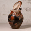 A handmade ceramic lidded jar with a glossy black body, orange-brown mottled glaze, and a light beige interior, set against a textured off-white wall.