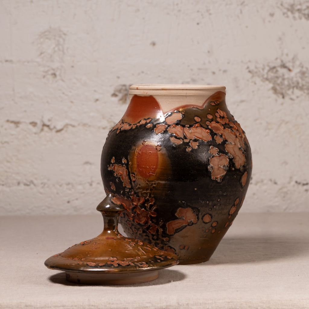 A handmade ceramic lidded jar with a glossy dark brown and black glaze, accented with coppery orange patches and a cream-colored rim, set against a textured light gray wall.